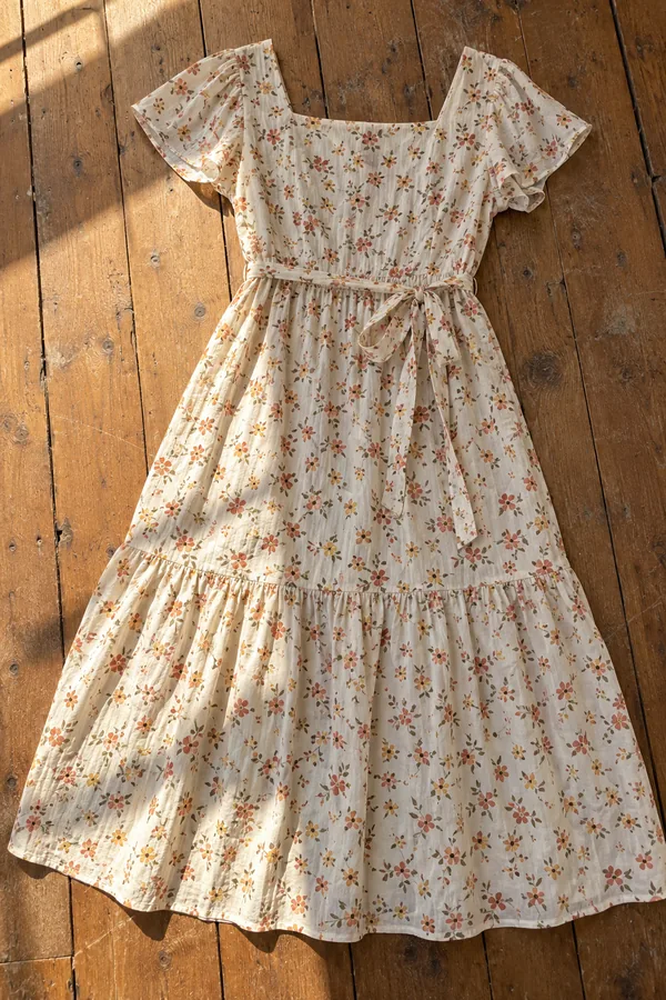 Cream cotton midi dress with a small terracotta and mustard floral print, flutter sleeves and a tie belt, laid flat on sunlit wooden floorboards.