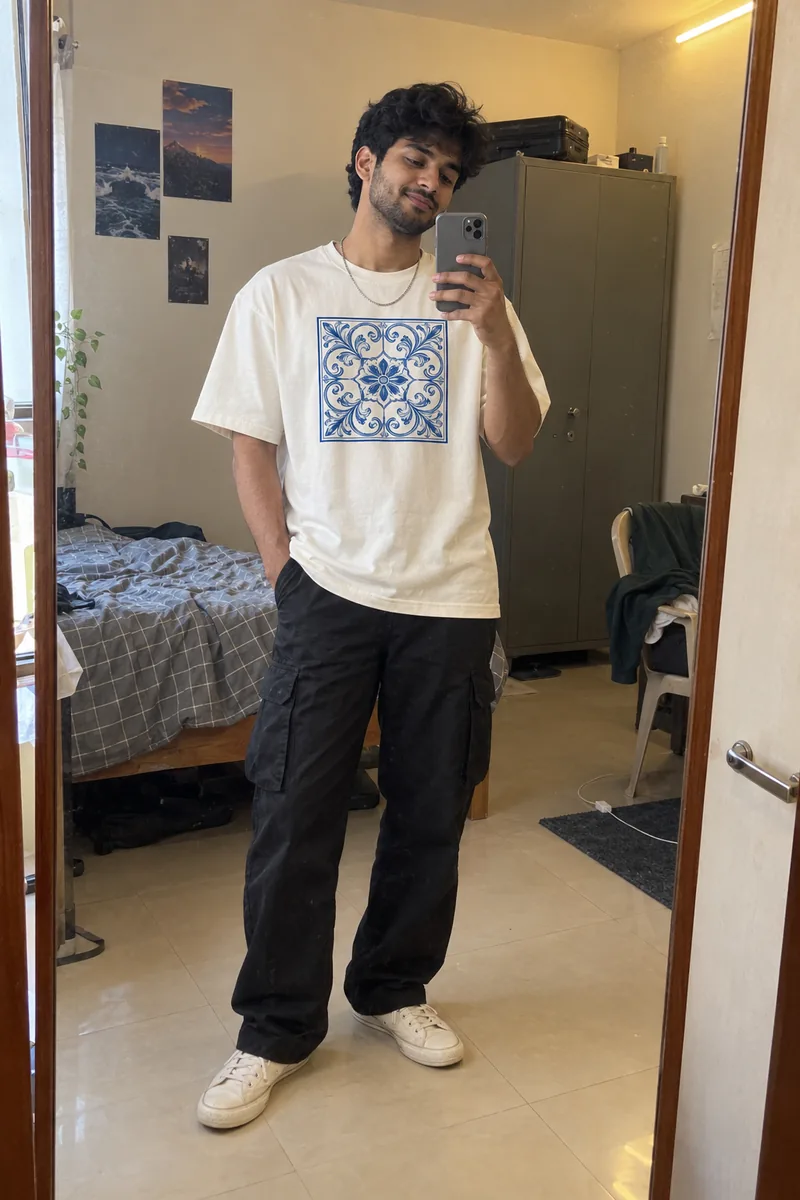 Young Indian man taking a mirror selfie in a lived-in bedroom, wearing the off-white tile-print tee oversized with black cargo trousers and white canvas sneakers.