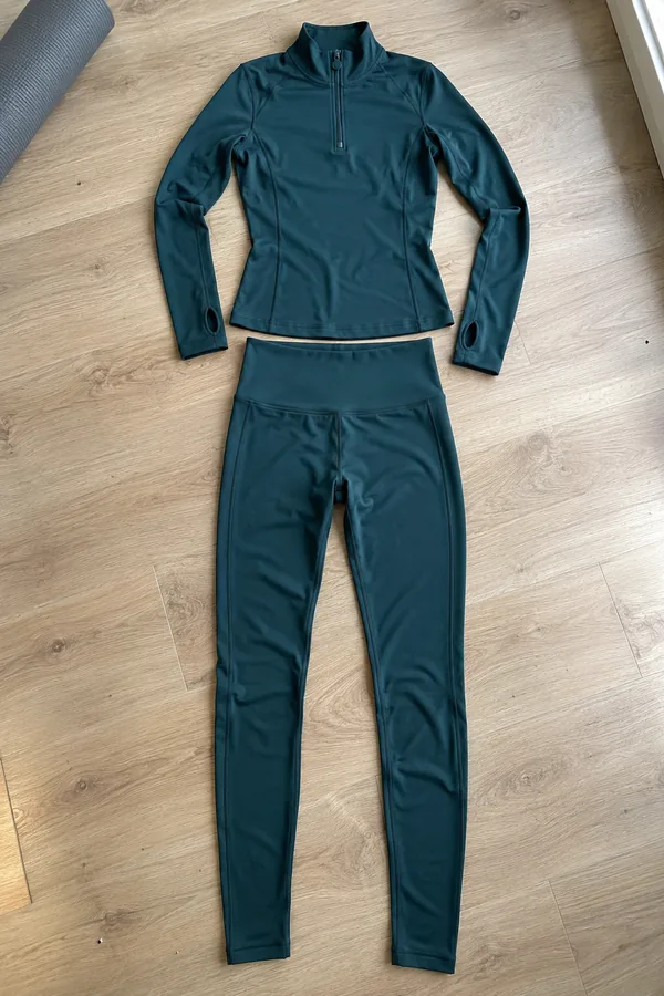Deep teal half-zip long-sleeve training top and matching high-waist leggings laid flat on a light wood floor next to a rolled yoga mat.